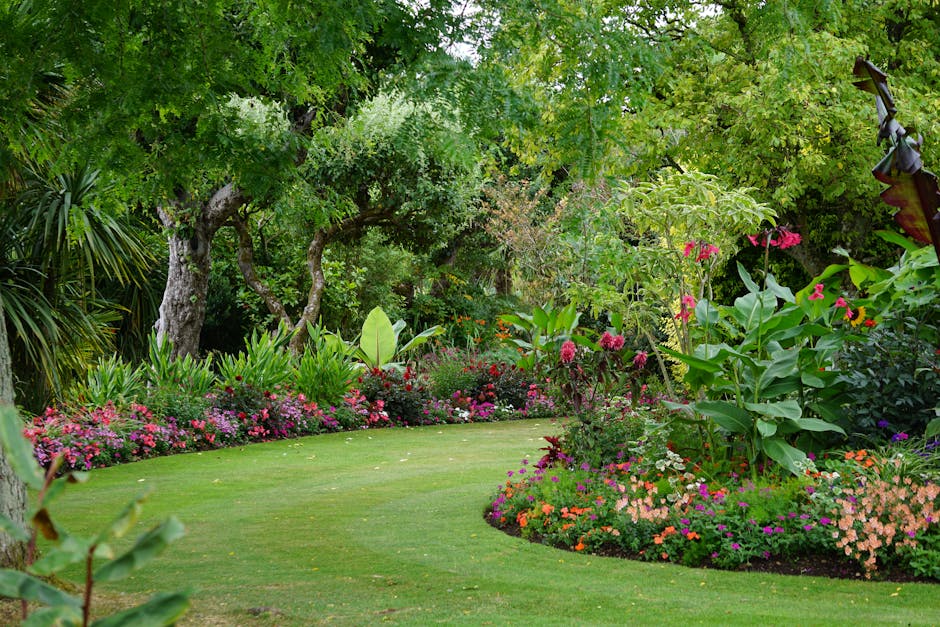 A lush, well-maintained garden scene featuring a neatly trimmed lawn interwoven with vibrant flower beds and borders. The foreground displays a gently curved, evenly cut grassy area bordered by colorf